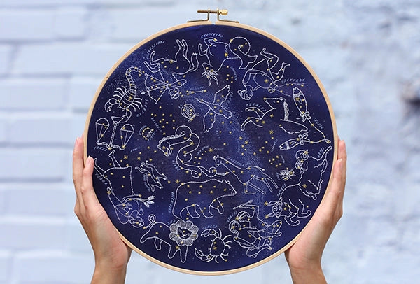 Embroidery hoop with zodiac constellation design held by hands against a blurred background